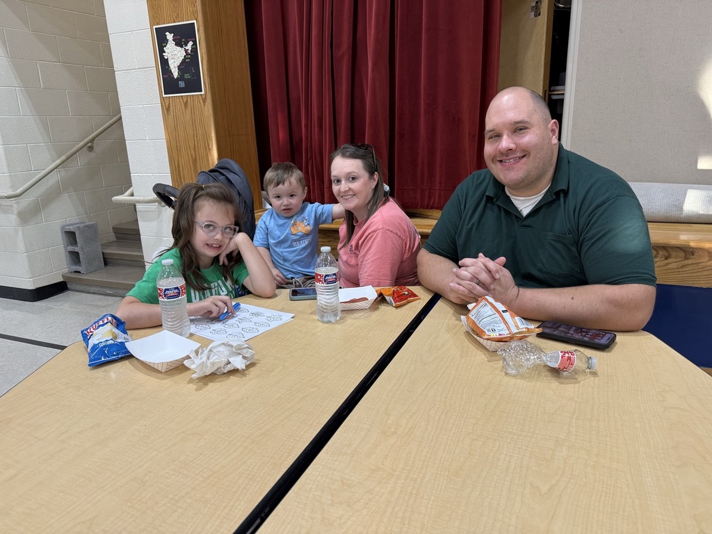 A family of four smiles for a photo while seated at a table during a Coalition for Kids (C4K) Family Night. Snacks, water bottles, and a coloring activity are spread across the table.
