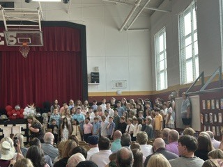 A large group of students stands on risers in a school gymnasium during an awards ceremony. Parents and family members are seated in the foreground facing the students.