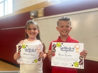 Two students stand side by side in a school gymnasium, each holding a Certificate of Recognition.
