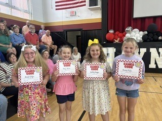 Four students stand in a row in a school gymnasium, each holding a "Game Changer" certificate. Red and white balloon decorations and a seated audience are visible in the background.