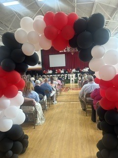A balloon arch made of black, white, and red balloons frames the entrance to a school gymnasium set up for an awards ceremony. Rows of chairs filled with attendees face a stage with a projected screen displaying "HAYNESFIELD" in the background.