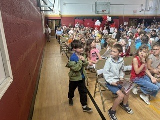 A young student stands in a school gymnasium aisle holding a certificate, with rows of seated students visible in the background alongside red and white balloon decorations.