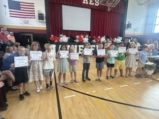 Eight students stand in a row in a school gymnasium, each holding a certificate. Large marquee letters spelling "HAYNESFIELD" are visible behind them, along with red and white balloon decorations and an audience in the background.