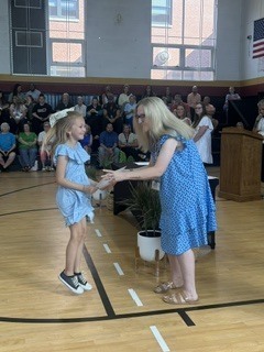 An adult woman in a blue floral dress bends toward a young girl in a blue outfit during what appears to be an awards presentation in a school gymnasium. A large audience is seated in the background.