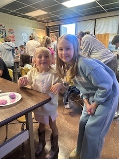 Two children smile while standing beside a cafeteria table covered with art supplies. Other students and adults are visible in the background.