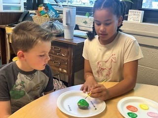 Two students sit at a round table. One paints a green rock while the other watches. A window and classroom supplies are visible in the background.