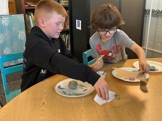 Two boys sit at a round table painting rocks. One wears glasses and holds a paintbrush while the other wipes his hands with a napkin.