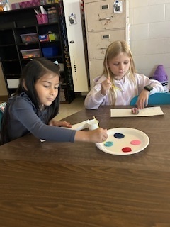 Two young students paint rocks at a classroom table using a plate of primary-colored paint. A cabinet and classroom posters are in the background.
