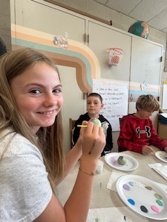 A smiling student holds a small rock while painting. Behind her, classmates paint at desks with plates of colorful paint.
