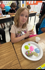 A student sits at a desk painting a rock with bright colors including pink, gold, and blue. A cup of water and paint plate are on the desk.