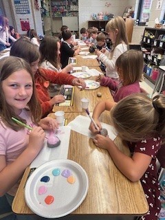 Several students sit in rows of desks painting rocks. A girl in a pink shirt smiles at the camera while others focus on their artwork.