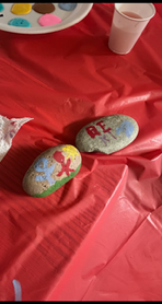 Two painted rocks on a red tablecloth—one with a red stick figure and the other with red letters “RE” and blue sky-like background.
