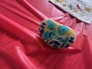 A smooth rock painted with teal and dark blue shapes on a yellow background rests on a red plastic tablecloth.