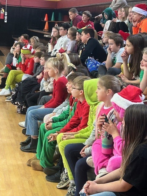 Group of elementary students seated on gym bleachers watching a presentation, some wearing holiday clothing.