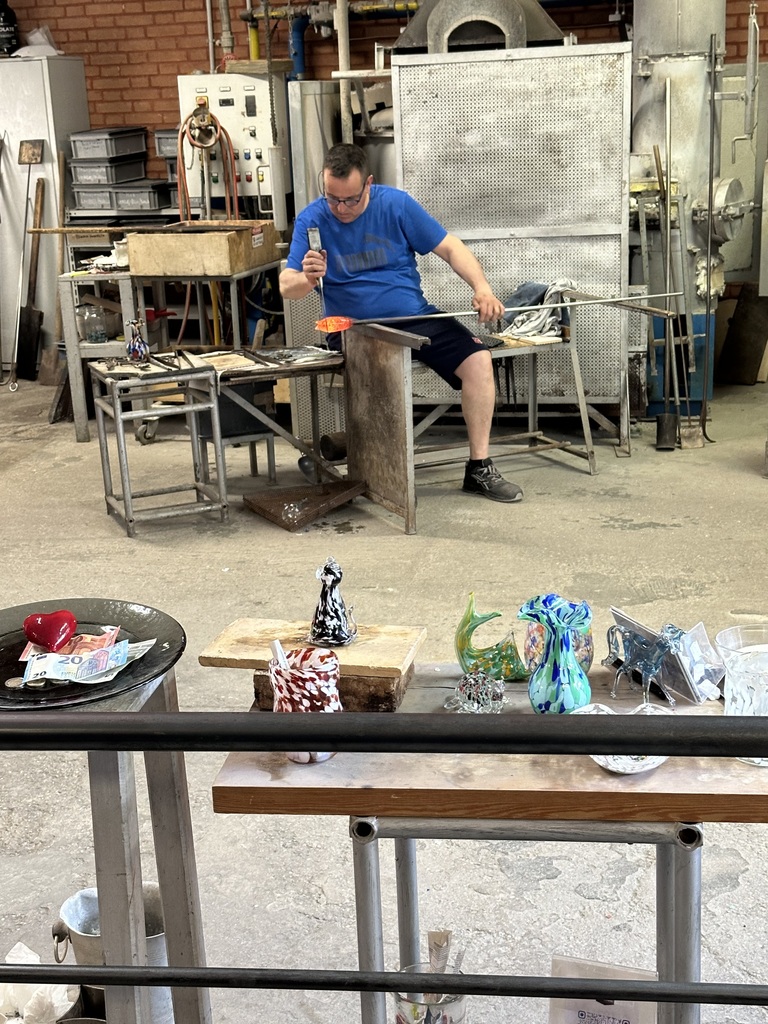 A man in casual clothes working on a molten-hot glass sculpture, with many colorful sculptures in the shape of flowers, vases, and cats in the foreground