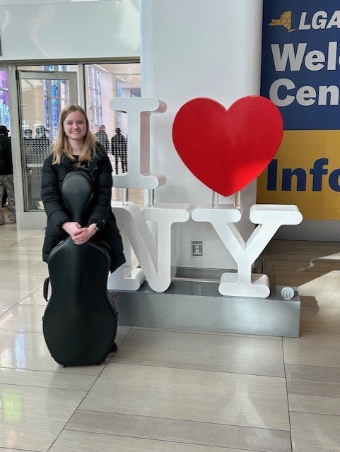 Elizabeth Biever standing in front of the iconic " I love NY" sign with her cello case.