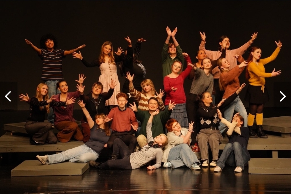 Image of multiple students on stage with arms outstretched in all directions with various emotions on their faces.