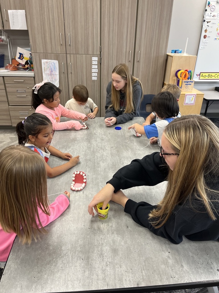 Preschool students around a table while an older student leads a dental health lesson.
