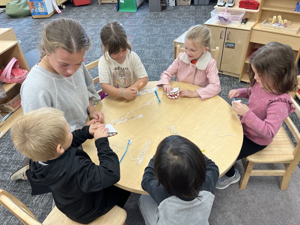 Preschool students around a table while an older student leads a dental health lesson.