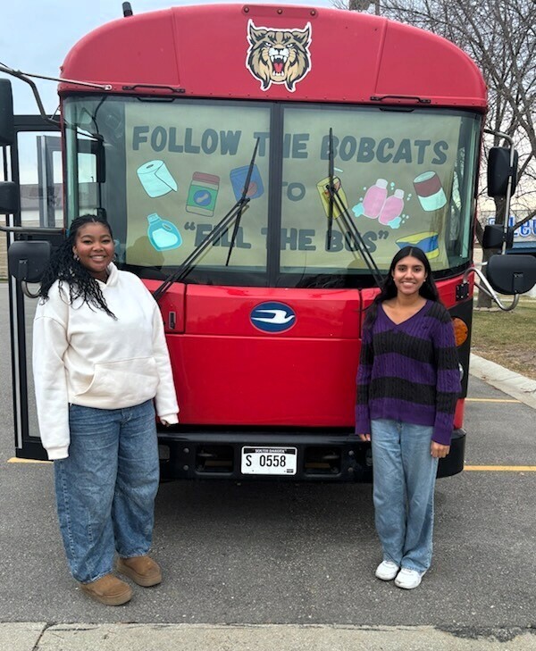 Two students standing in front of the red, bobcat bus. 