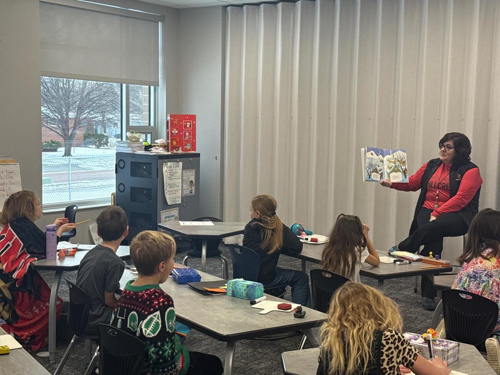 Principal Mogard holding a book she is reading in front of a class of students. A student is seen engaging with the story and seems to be asking a question.