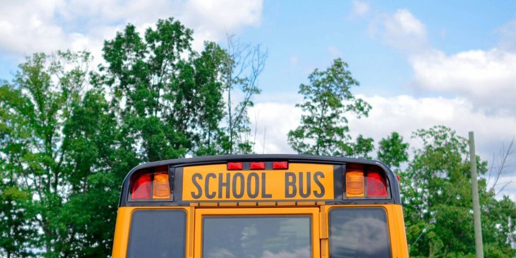 Back of a yellow school bus against a background of trees and blue sky with some clouds.