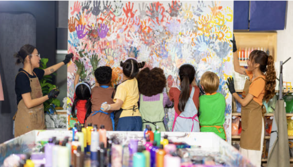 A group of children, wearing aprons and gloves, are painting on a large canvas with a teacher assistants.