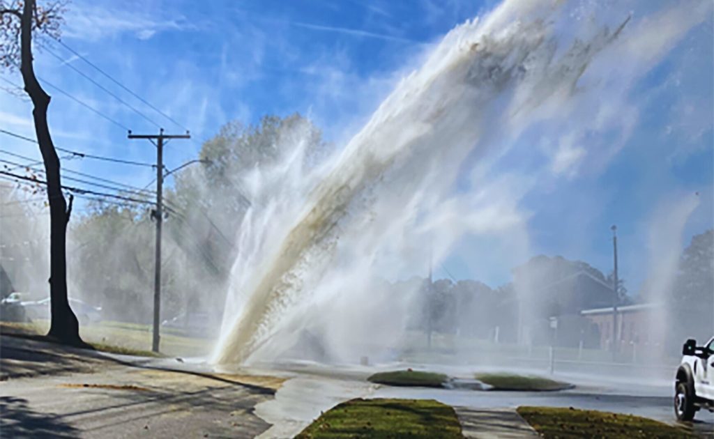 A water main break and a broken hydrant spraying water into the air, creating a fountain effect. Behind it, a tree, a utility pole, and a house.
