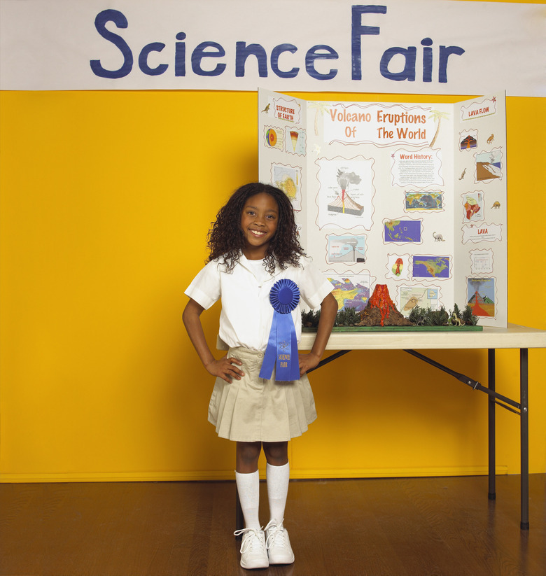 A young girl with a blue ribbon stands beside a science fair project table. The backdrop is a yellow wall with "Science Fair" in blue.