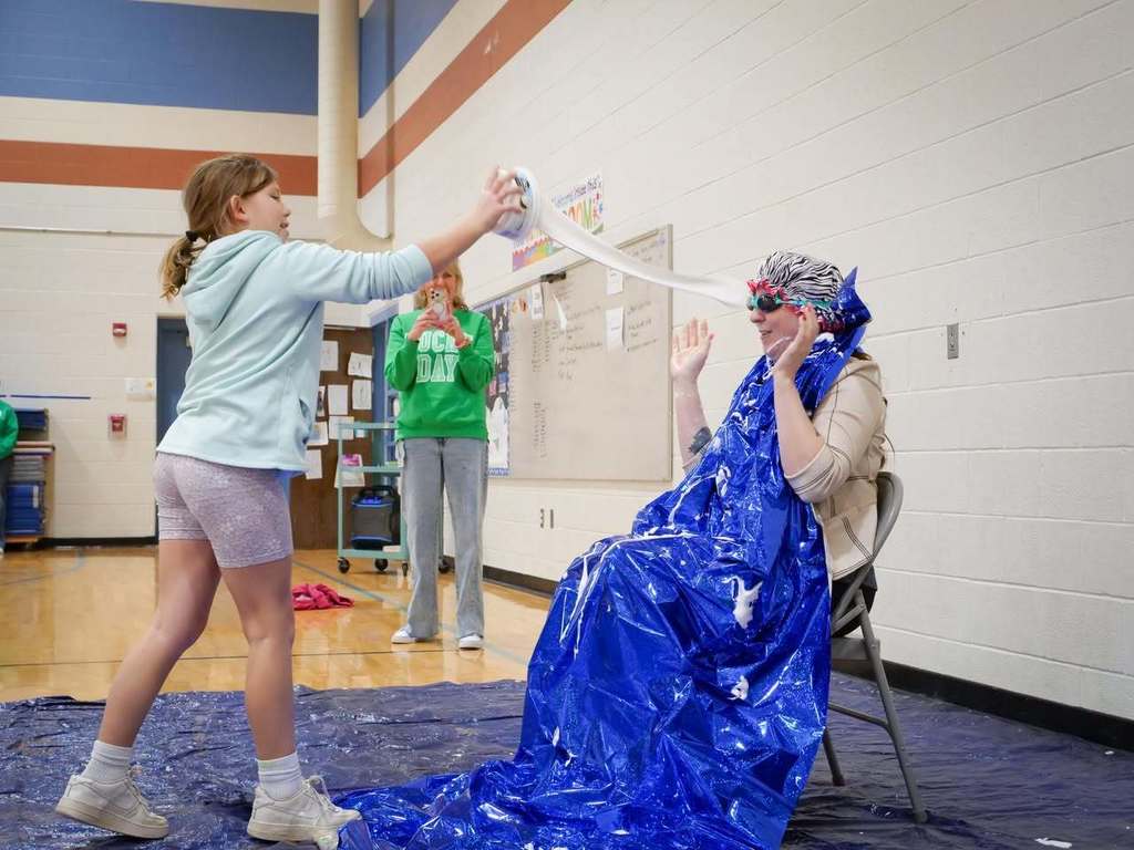 Two individuals in a gym; one holding a bubble wand, blowing bubbles towards another in a blue cape.