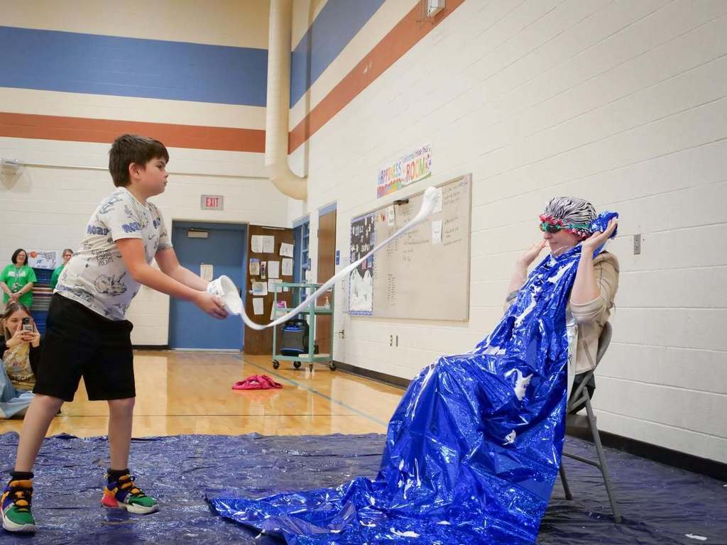 A boy in a gym holding a long blue plastic sheet, pulling it towards a seated person.