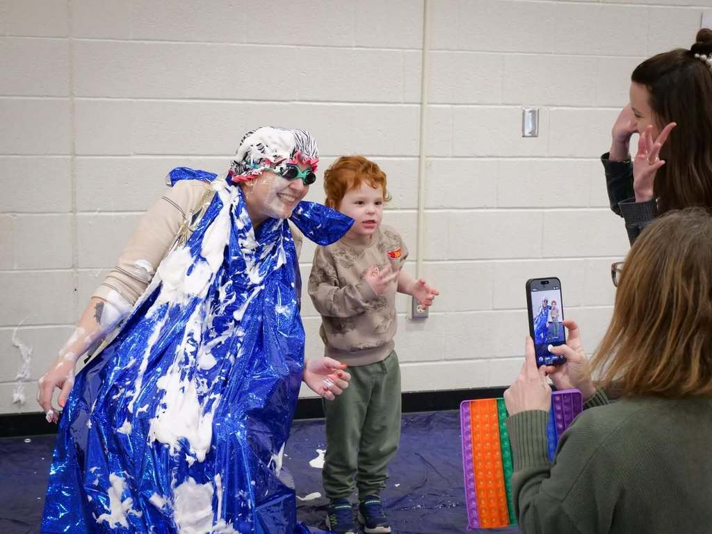 An adult with blue paint on their clothing dances with a young child in a room. A woman takes a photo.