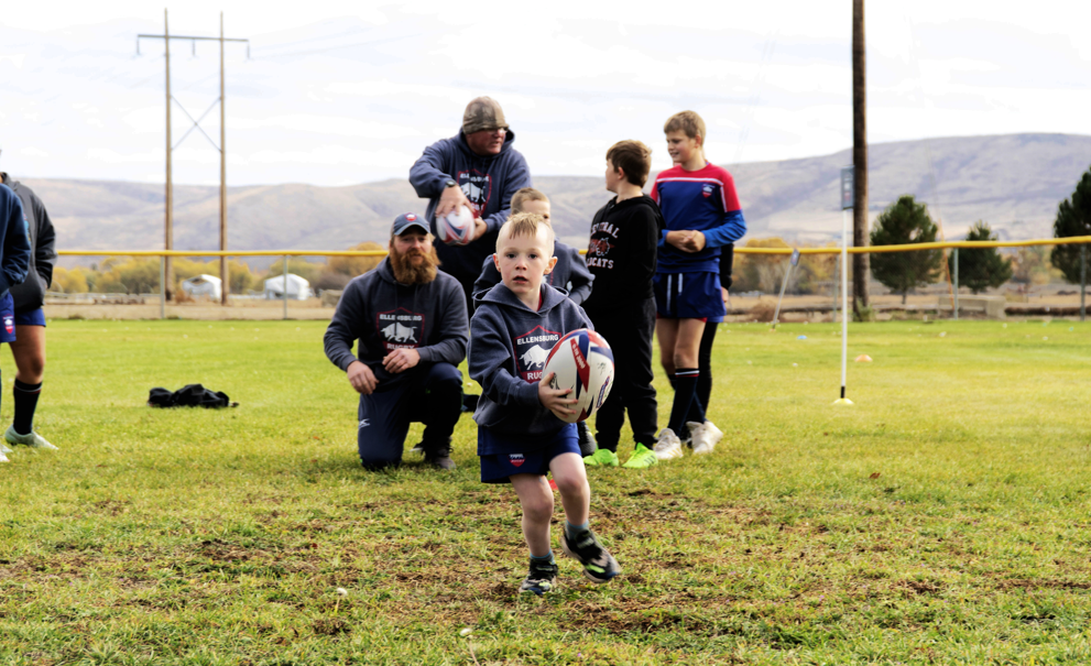 Several people on a field, with one child holding a rugby ball, running while others watch.