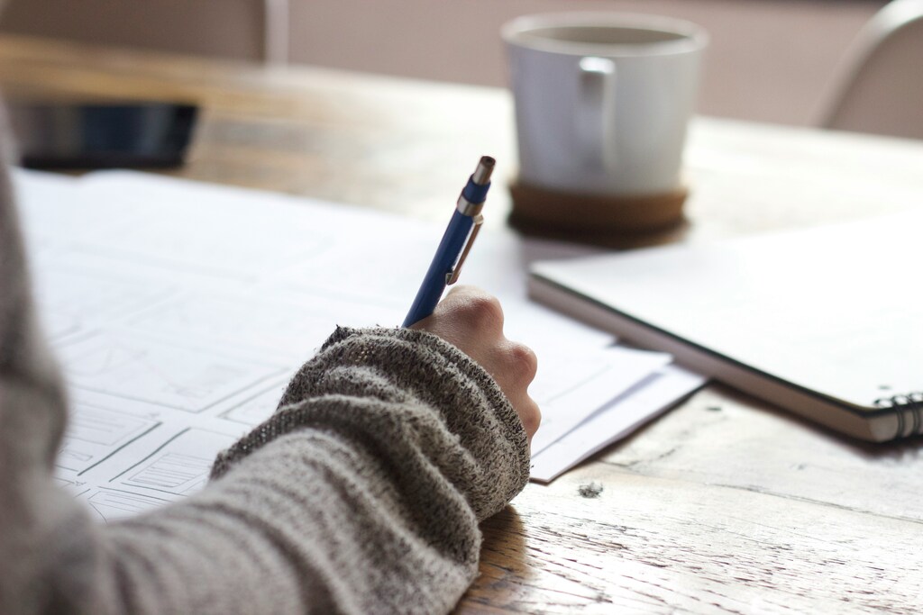 Person seated at a wooden table, writing on paper with a pen, surrounded by notebooks and a cup.
