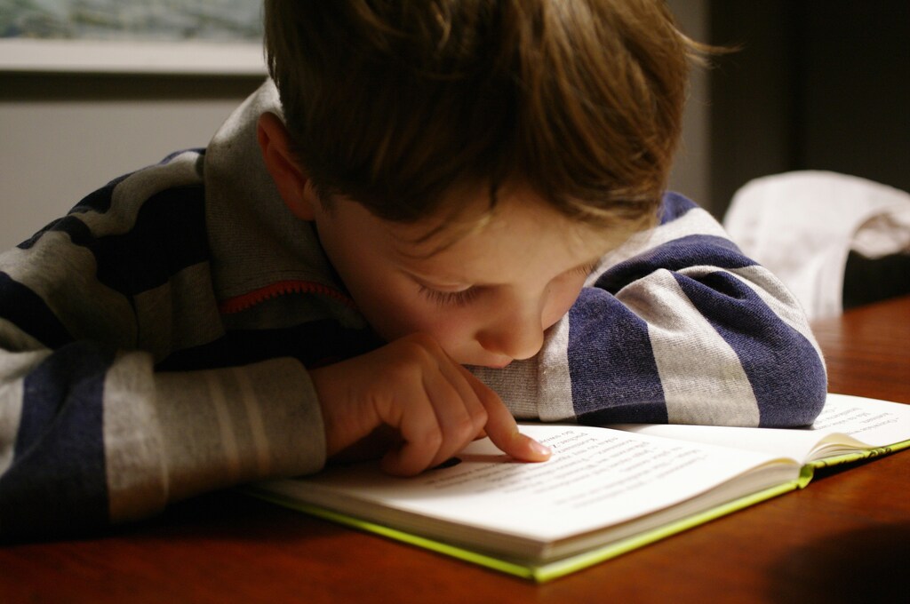 A child in a striped shirt reads a book at a table, head down, focused on the text.