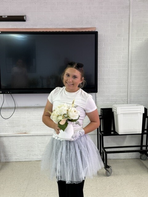 A person in a white shirt and tutu holds a bouquet of white flowers, standing in front of a TV.