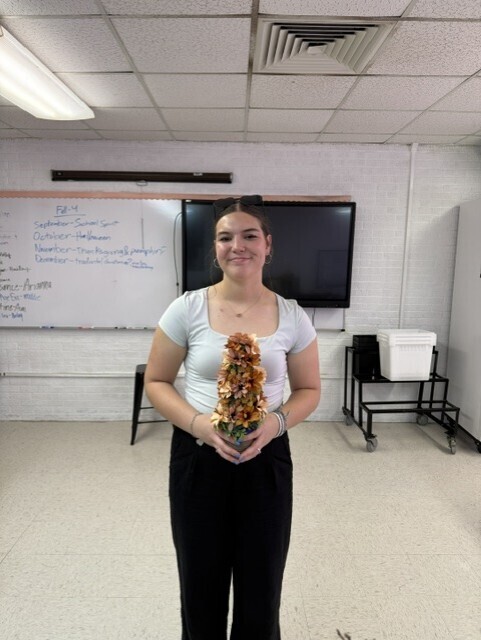 A woman in a classroom holds a tall, colorful object in her hands. Behind her is a whiteboard and a monitor.