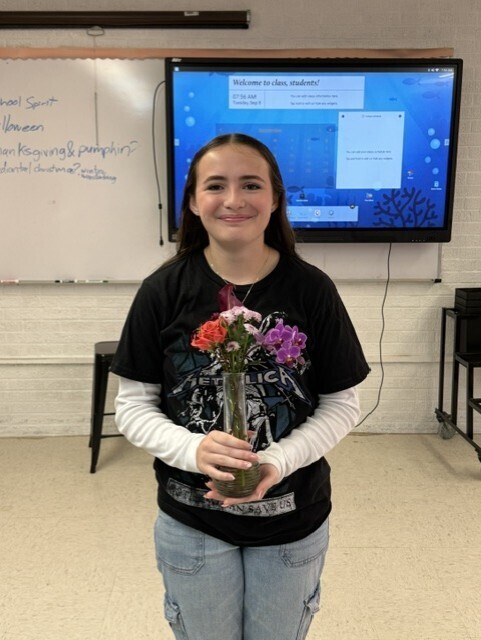 A woman in a black t-shirt holds a vase with flowers in a room with a whiteboard.