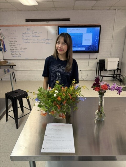 Woman stands at a table with flowers, a whiteboard in the background, and a television monitor.