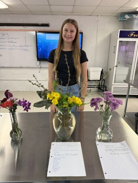 A young person stands by a table with three vases of flowers. Behind them, a whiteboard and a TV are visible.