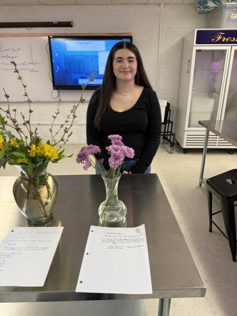 A woman stands at a table with flowers in vases. Papers are on the table. A monitor and whiteboard are in the background.