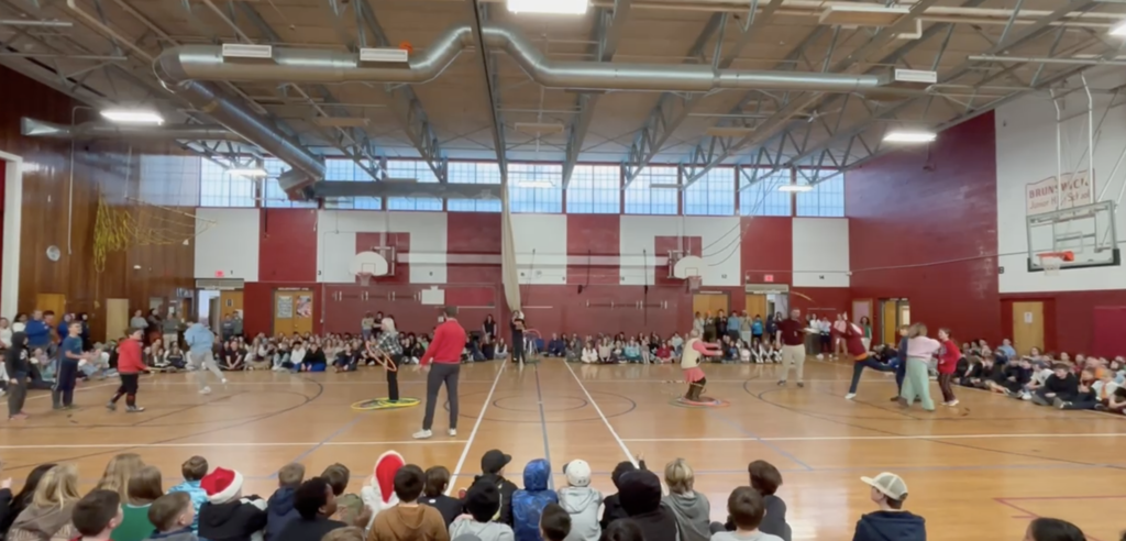 Group of students seated on gym floor while students are tossing hula hoops at a staff member.