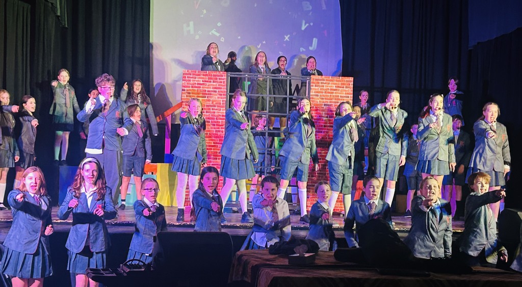 Cast of Matilda standing on stage poitning at audience. 
