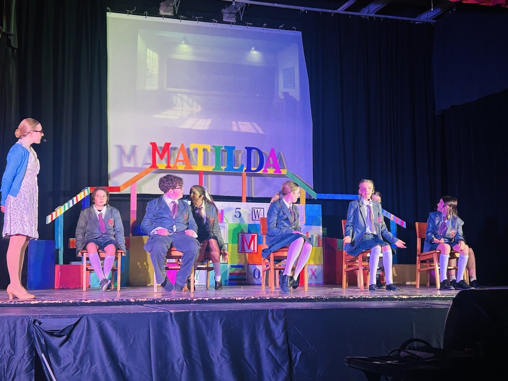 Cast of Matilda sitting in a classroom on stage with a teacher at the front of the room.