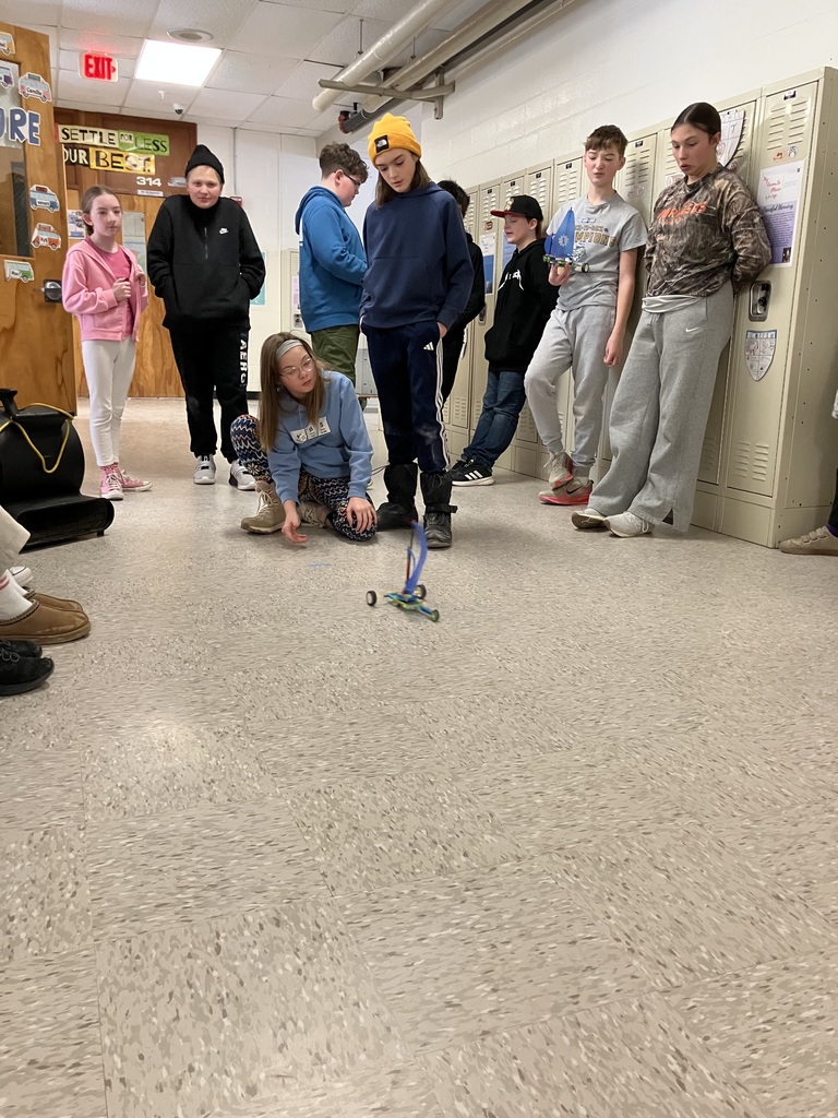 Students stand in a school hallway while one student crouches on the floor, guiding a LEGO sailboat that moves down the hallway using wind power.
