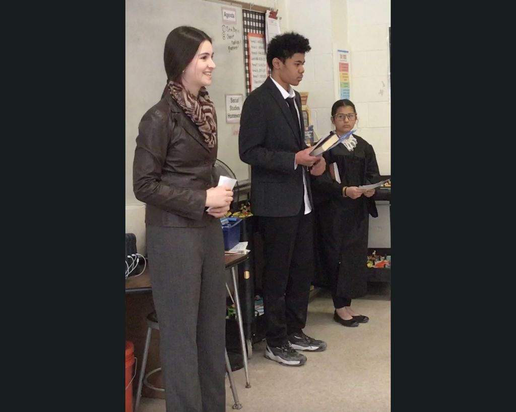 Three students standing in the front of a class dressed in costume as influential American figures.