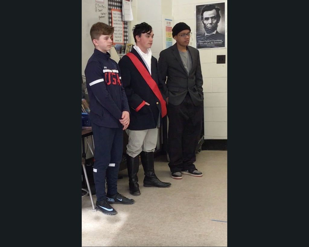 Three students standing in the front of a class dressed in costume as influential American figures.