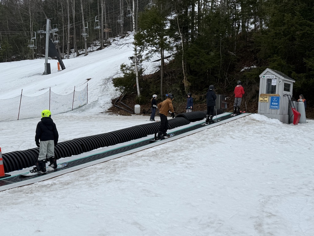 Students riding a carpet lift on skis up a small hill.