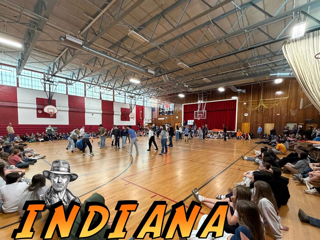 Students seated on the floor around the gym while a group of students stands in the center of the gym in a circle passing a ball around.