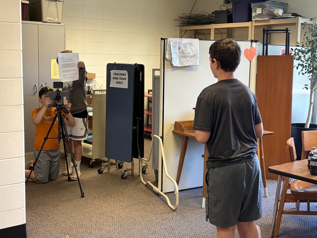Two students behind a camera on a tripod and one student standing infront of the camera filming a scene. 